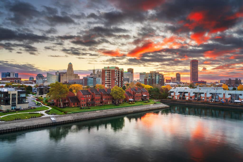 Die Skyline von Buffalo, New York, bei Sonnenuntergang. Die Aufnahme zeigt die Stadtansicht vom Ufer eines Flusses oder Kanals aus, an dem sich eine Reihe von Backsteinhäusern befinden. Die tief hängenden Wolken am Himmel sind in lebhaften Rot- und Orangetönen gefärbt und spiegeln sich auf der Wasseroberfläche.