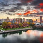 Die Skyline von Buffalo, New York, bei Sonnenuntergang. Die Aufnahme zeigt die Stadtansicht vom Ufer eines Flusses oder Kanals aus, an dem sich eine Reihe von Backsteinhäusern befinden. Die tief hängenden Wolken am Himmel sind in lebhaften Rot- und Orangetönen gefärbt und spiegeln sich auf der Wasseroberfläche.