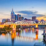 Skyline von Nashville, Tennessee, bei Sonnenuntergang mit beleuchteter Brücke über den Cumberland River und modernen Hochhäusern im Hintergrund.