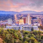 Abendstimmung in Salt Lake City Das Bild zeigt die Skyline von Salt Lake City im Abendlicht. Im Vordergrund steht das markante Utah State Capitol mit seiner großen Kuppel, umgeben von Bäumen und Grünflächen. Dahinter erstreckt sich die moderne Innenstadt mit zahlreichen Hochhäusern und Gebäuden, die sich bis zu den Ausläufern der Rocky Mountains erstreckt. Der Himmel ist in weichen Rosa- und Lilatönen gefärbt, was der Szene eine stimmungsvolle Atmosphäre verleiht.