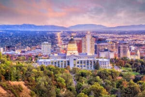 Abendstimmung in Salt Lake City Das Bild zeigt die Skyline von Salt Lake City im Abendlicht. Im Vordergrund steht das markante Utah State Capitol mit seiner großen Kuppel, umgeben von Bäumen und Grünflächen. Dahinter erstreckt sich die moderne Innenstadt mit zahlreichen Hochhäusern und Gebäuden, die sich bis zu den Ausläufern der Rocky Mountains erstreckt. Der Himmel ist in weichen Rosa- und Lilatönen gefärbt, was der Szene eine stimmungsvolle Atmosphäre verleiht.