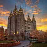 Der Salt-Lake-Tempel in Salt Lake City bei Sonnenuntergang. Das große, gotische Gebäude ist hell beleuchtet und steht vor einem dramatischen Himmel mit orangefarbenen und grauen Wolken. Die Bäume im Vordergrund sind mit Lichtern geschmückt.