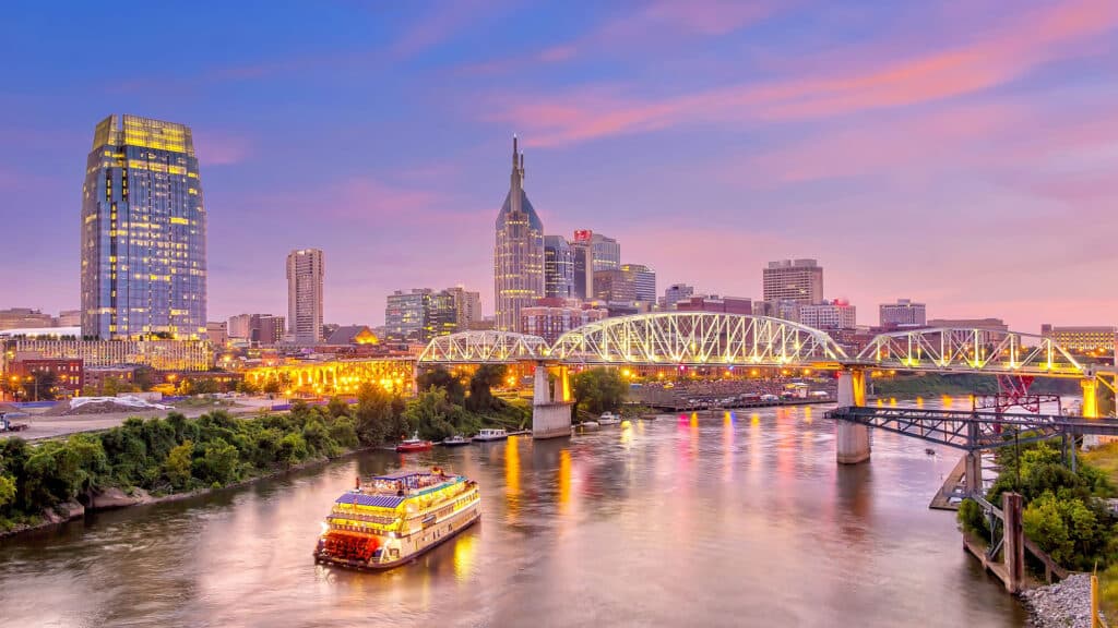 Die beleuchtete Skyline von Nashville, Tennessee, bei Sonnenuntergang. Ein Raddampfer-Schiff fährt auf dem Cumberland River im Vordergrund. Die Shelby Street Pedestrian Bridge führt über den Fluss und verbindet das Stadtzentrum mit den östlichen Stadtteilen.