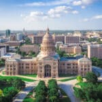 Das ist das Texas State Capitol in Austin, Texas, aufgenommen aus der Vogelperspektive. Das Gebäude ist von einer gepflegten Grünanlage umgeben und die Skyline der Stadt Austin ist im Hintergrund zu sehen. Der Himmel ist blau mit einigen weißen Wolken.