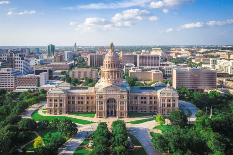 Das ist das Texas State Capitol in Austin, Texas, aufgenommen aus der Vogelperspektive. Das Gebäude ist von einer gepflegten Grünanlage umgeben und die Skyline der Stadt Austin ist im Hintergrund zu sehen. Der Himmel ist blau mit einigen weißen Wolken.