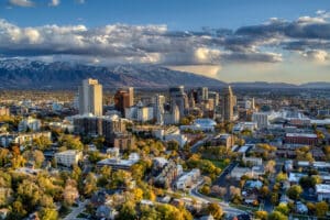 Die Skyline von Salt Lake City, Utah, bei Sonnenuntergang. Die Stadt liegt in einem Tal, umgeben von schneebedeckten Bergen. Die Bäume im Vordergrund zeigen Herbstfarben.