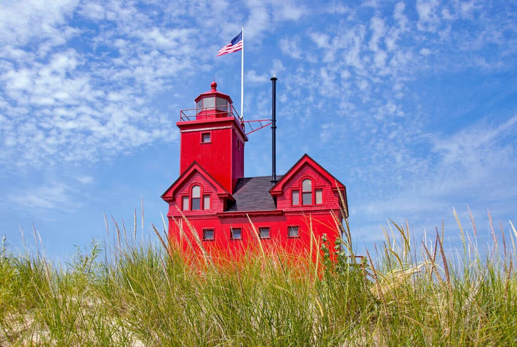 Roter Leuchtturm (Big Red) in Holland, Michigan, auf einer Düne mit hohem Gras, mit amerikanischer Flagge und blauem, bewölktem Himmel.