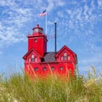 Roter Leuchtturm (Big Red) in Holland, Michigan, auf einer Düne mit hohem Gras, mit amerikanischer Flagge und blauem, bewölktem Himmel.